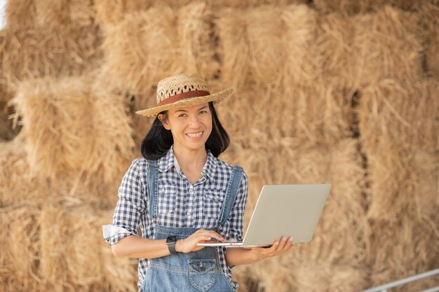 Asia joven agricultora con sombrero de pie en el campo y escribiendo en el teclado de la computadora portátil. Mujer con laptop supervisando el trabajo en tierras de cultivo, concepto de ecología, transporte, aire limpio, alimentos, productos biológicos