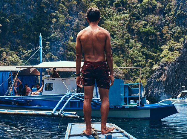 Un asiático sin camisa parado en la proa del barco en el fondo de un hermoso paisaje de bahía rocosa.