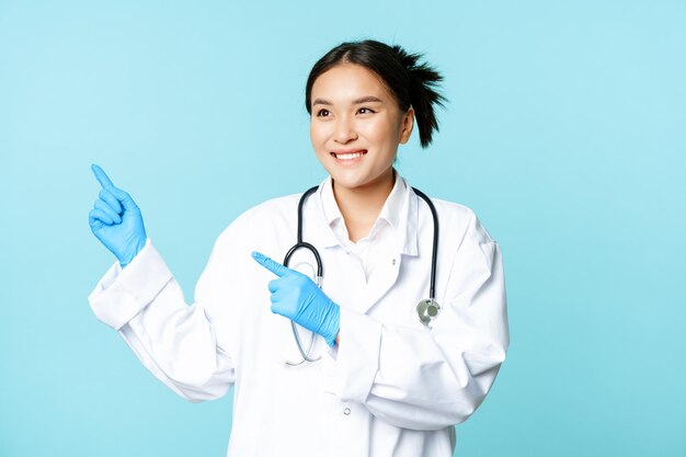 Asiático sonriente doctor médico trabajador de la salud señalando con el dedo a la izquierda mirando complacido en la publicidad ...