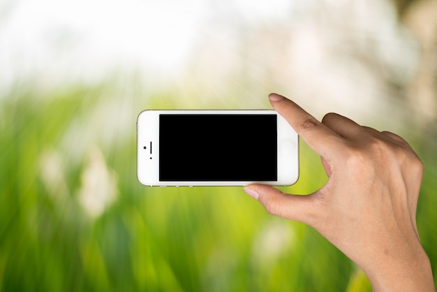 Asimiento de la mano de la mujer y teléfono elegante en luz del día con el fondo borroso verde de la naturaleza.