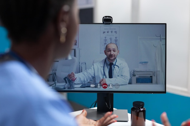 Asisstant afroamericano discutiendo los síntomas de la gripe con el médico a distancia durante la conferencia de la reunión de videollamada en línea que trabaja en la oficina del hospital. Llamada de telemedicina en la pantalla de la computadora