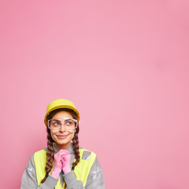 Asistente de constructor de mujer mantiene las manos juntas mira arriba pensativamente piensa en la construcción del nuevo edificio usa gafas de seguridad de casco protector y uniforme aislado sobre pared rosa.