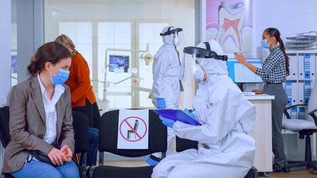 Asistente de dentista con equipo de ppe hablando con el paciente antes de la consulta durante la epidemia de coronavirus sentado en sillas en el área de espera manteniendo la distancia. Concepto de nueva visita al dentista normal.