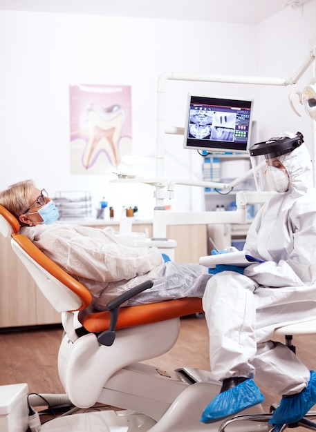 Asistente de dentista con traje de materiales peligrosos contra el coronavirus sin hablar con el paciente mayor. Anciana en uniforme protector durante el examen médico en la clínica dental.