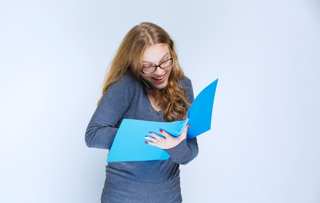 Asistente hablando con el teléfono blanco revisando la carpeta azul y tomando notas, y se ve muy feliz.