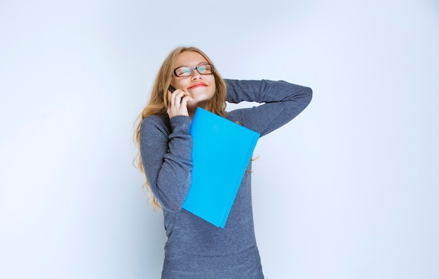 Asistente hablando con el teléfono blanco revisando la carpeta azul y tomando notas, y se ve muy feliz.