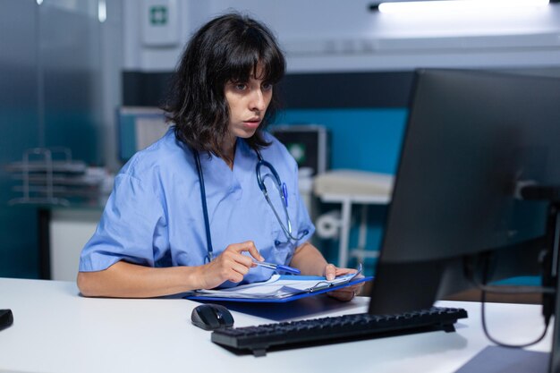 Asistente médico analizando documentos y archivos en el monitor por la noche. Enfermera mirando la computadora y trabajando para una cita médica con papeles de chequeo, haciendo horas extras en el escritorio.