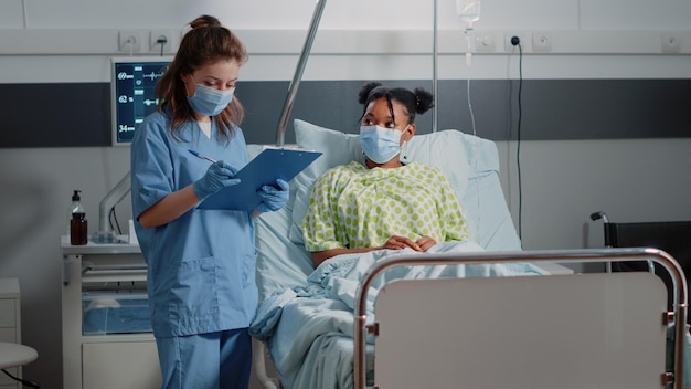 Foto gratuita asistente médico consultando a un paciente enfermo, usando mascarillas en la sala del hospital. enfermera discutiendo con una joven sobre el tratamiento de la salud y tomando notas durante la pandemia