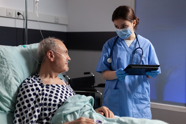 Asistente médico con máscara de protección que controla el tratamiento del hombre mayor leyendo notas en tablet pc