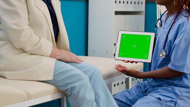 Asistente médico sosteniendo una tableta digital con pantalla verde, haciendo una visita de control con un paciente mayor. Enfermera y mujer analizando plantilla cromakey aislada con fondo de maqueta y copyspace.