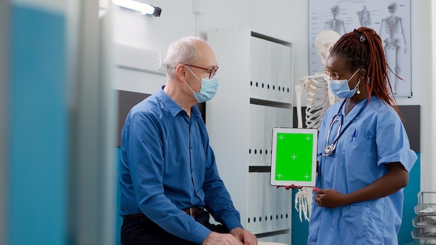 Asistente que muestra una tableta con pantalla verde a un paciente mayor durante la pandemia de covid 19. mirando la plantilla de maqueta aislada con copyspace en blanco y pantalla cromakey en la visita de control.