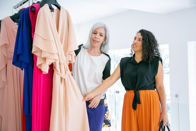 Asistente de tienda ayudando al cliente a elegir la tela. Los compradores tocaron un vestido nuevo colgado de un estante. Tiro medio. Tienda de moda o concepto minorista.