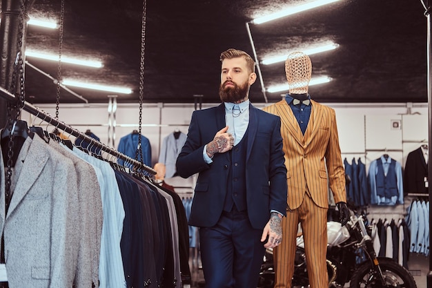 Asistente de tienda barbudo elegantemente vestido con pie cerca de maniquí en una tienda de ropa masculina.