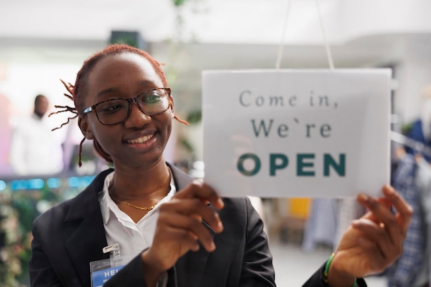 Foto gratuita la asistente de la tienda de ropa sonriente afroamericana cuelga un cartel de apertura en la puerta de entrada. el trabajador de la tienda de moda alegre pone un cartel en la ventana notando el comienzo del día de trabajo