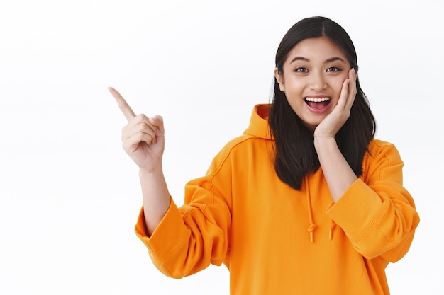 Asombrada chica asiática feliz encontró la mejor oferta increíble mostrándote, señalando con el dedo la esquina superior izquierda en el anuncio y sonriendo asombrada, tocando la mejilla enrojecida y sorprendida, pared blanca