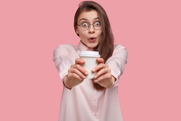 Asombrada hermosa joven con mirada sorprendida estira las manos con café para llevar, vestida con camisa formal, mantiene la mandíbula caída, vestida elegantemente