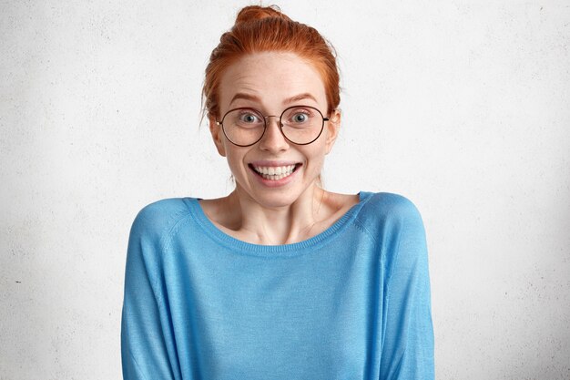 Asombrada hermosa modelo femenina llena de alegría con cabello rojo y piel pecosa, viste un suéter azul casual, mira a través de gafas redondas sorprendentemente