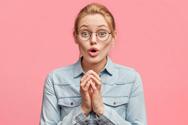 Asombrada hermosa modelo de mujer joven rubia con ojos azules se ve con desconcierto, queda atónita al ver algo inesperado, usa gafas redondas y ropa de mezclilla de moda.