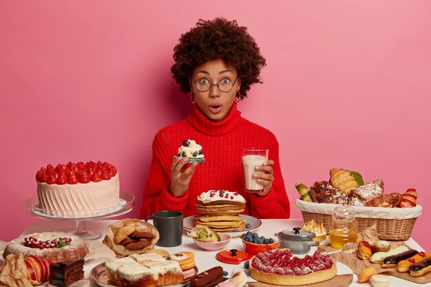 Asombrada joven mujer afro disfruta comiendo deliciosos cupcakes con yogur, disfruta de una cena festiva, sorprendida por la cantidad de calorías que comió, usa un suéter rojo, sabe un postre cremoso