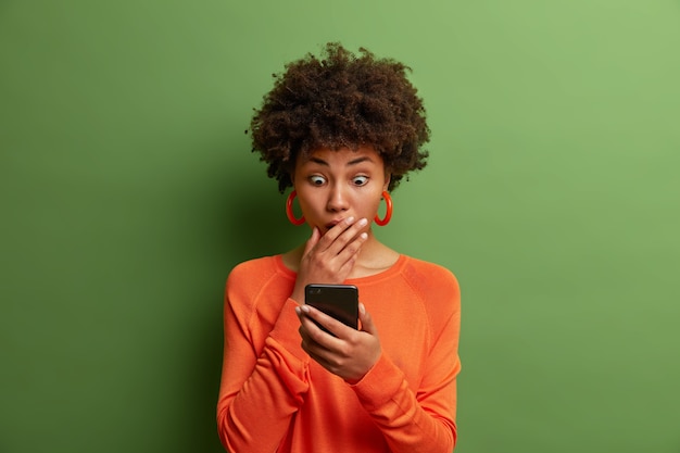 asombrada mujer adulta profundamente sorprendida, mira fijamente la pantalla del teléfono inteligente, lee noticias impactantes en el sitio web, se viste con un jersey naranja, tiene los ojos saltones, se tapa la boca. ¡Dios mío, es horrible!