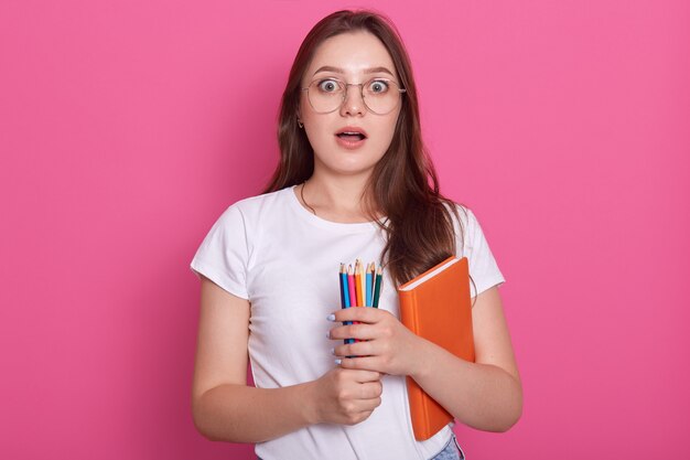 Asombrada mujer con la boca abierta, lleva un libro de texto y lápices de colores para escribir o dibujar, y está lista para tomar notas.