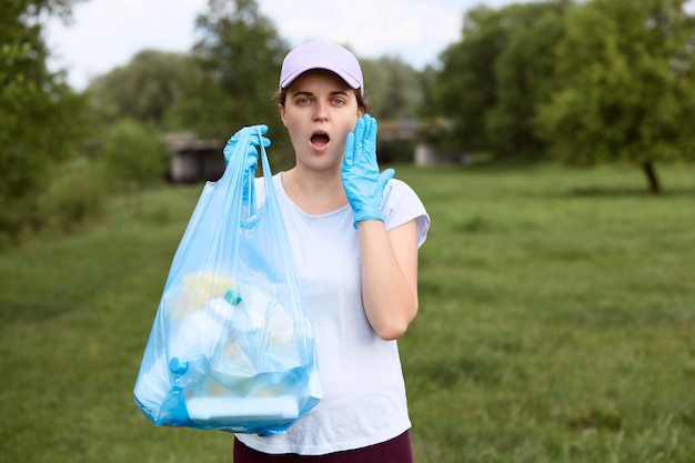 Asombrada niña con la boca abierta de pie, con la bolsa de basura en la mano, con la palma de la mano en la mejilla