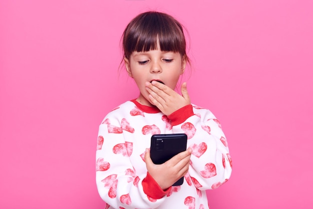 Asombrada niña vistiendo un puente casual mirando el teléfono inteligente con mirada de sorpresa, cubriendo la boca con la palma de la mano leyendo noticias impactantes, posando aislado sobre una pared rosa.