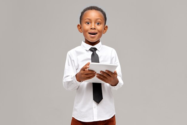 Asombrado niño de piel oscura con camisa blanca y corbata negra disfrutando de una conexión inalámbrica a Internet de alta velocidad en una tableta digital con una mirada sorprendida y asombrada, viendo dibujos animados en línea