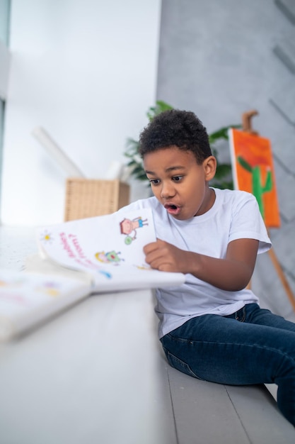 Asombro. Niño en edad escolar de piel oscura con camiseta blanca mirando sorprendido el libro con la boca abierta sentado en una habitación luminosa a la luz del día