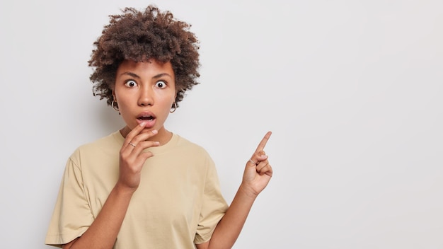 Asombrosa mujer hermosa con el pelo rizado muestra puntos de anuncio en el anuncio con expresión de asombro vestida con una camiseta beige casual aislada sobre fondo blanco Concepto de promoción