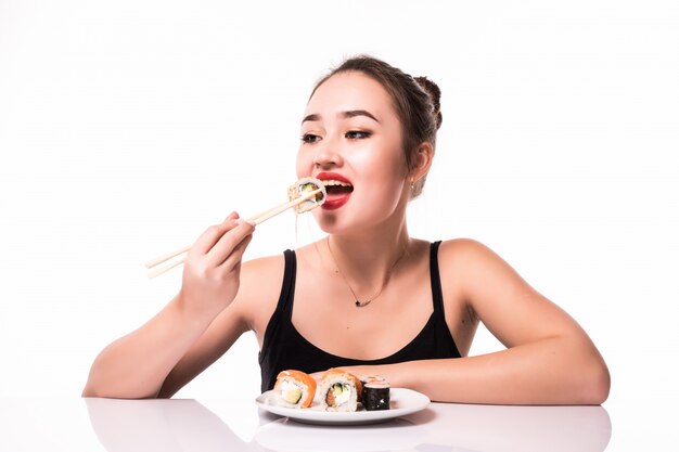 Aspecto bastante asiático con peinado modesto sentarse en la mesa comer rollos de sushi sonriendo aislado en blanco