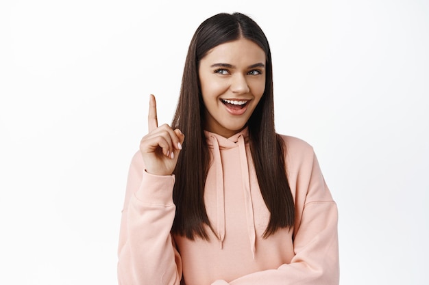 Astuta chica morena sonriendo y riendo, mirando a un lado pensativo y levantando el dedo, teniendo una idea, de pie pensativo con buen plan