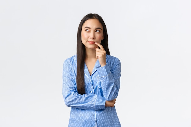 Astuta y pensativa chica asiática bonita en pijama azul, sonriendo y mirando en la esquina superior izquierda, imaginando algo, teniendo una idea interesante, de pie con fondo blanco en pijamas