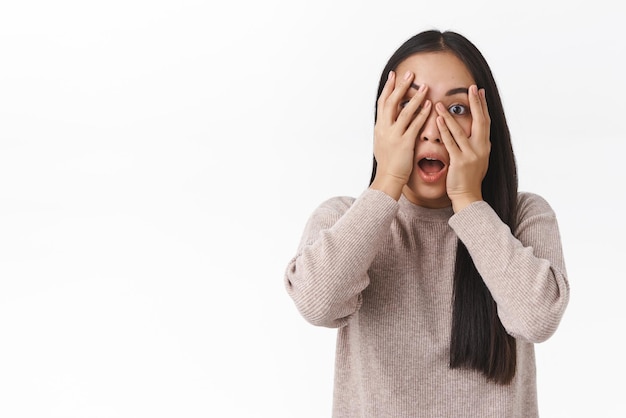 Foto gratuita asustada, avergonzada, linda chica asiática escondiendo la cara, tome las manos sobre los ojos y mirando a la cámara asustada o alarmada viendo una película de terror sola, payasos oscuros y asustados, de pie, con fondo blanco, sacudidos