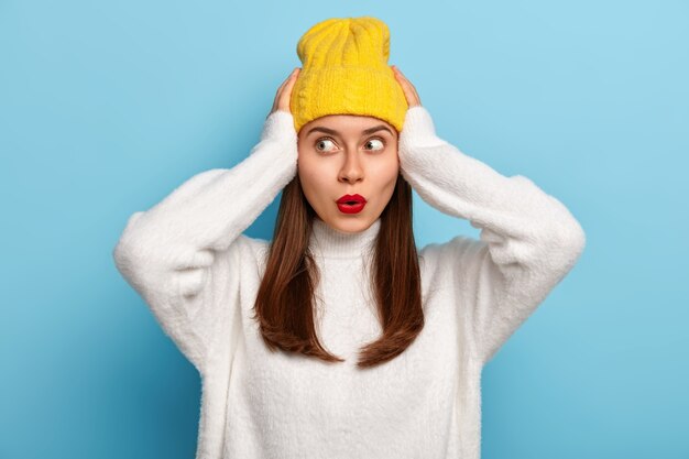 Asustada hermosa joven mantiene ambas manos en la cabeza, mira a un lado con terror, tiene el pelo largo y oscuro, usa un sombrero amarillo y un suéter blanco suave, enfocado a un lado, aislado sobre fondo azul