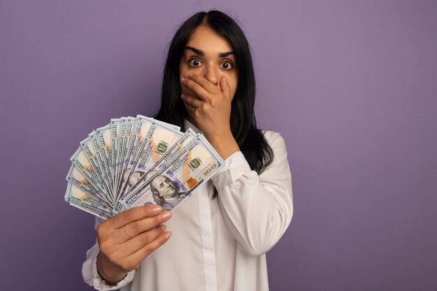 Asustada hermosa joven vestida con camiseta blanca sosteniendo dinero cubrió la boca con la mano aislada en púrpura con espacio de copia