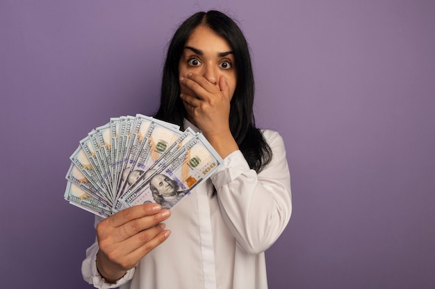 Asustada hermosa joven vestida con camiseta blanca sosteniendo dinero cubrió la boca con la mano aislada en púrpura con espacio de copia