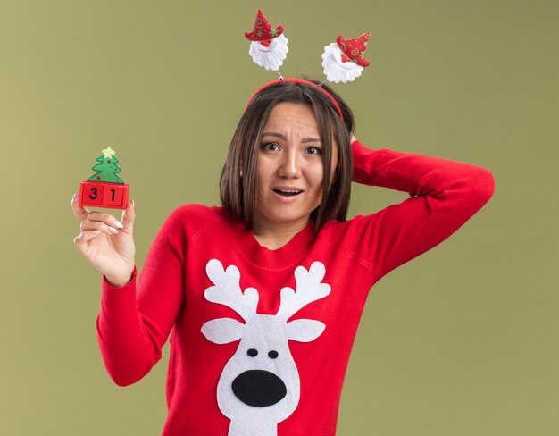 Asustada joven asiática vistiendo un aro de pelo de navidad sosteniendo un juguete de navidad poniendo la mano detrás de la cabeza aislada en la pared verde oliva