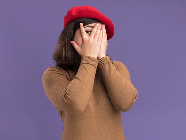 Asustada joven bastante caucásica con sombrero de boina cubre la cara con las manos a través de los dedos aislados en la pared púrpura con espacio de copia