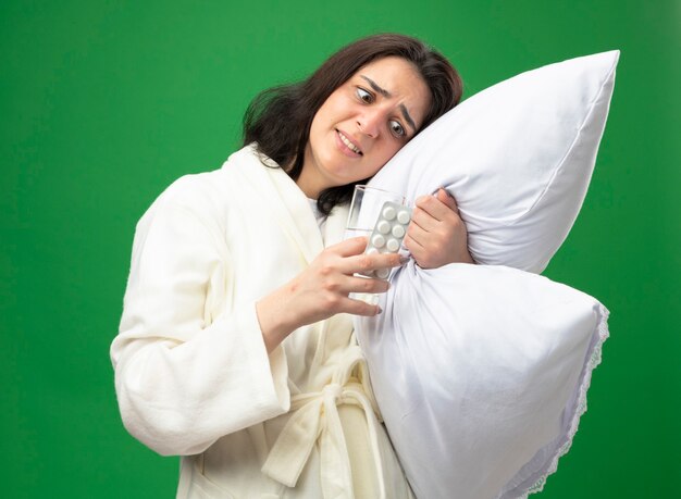 Asustada joven caucásica enferma vestida con bata abrazando la almohada poniendo la cabeza sobre ella sosteniendo un vaso de agua y un paquete de tabletas médicas mirándolos aislado sobre fondo verde