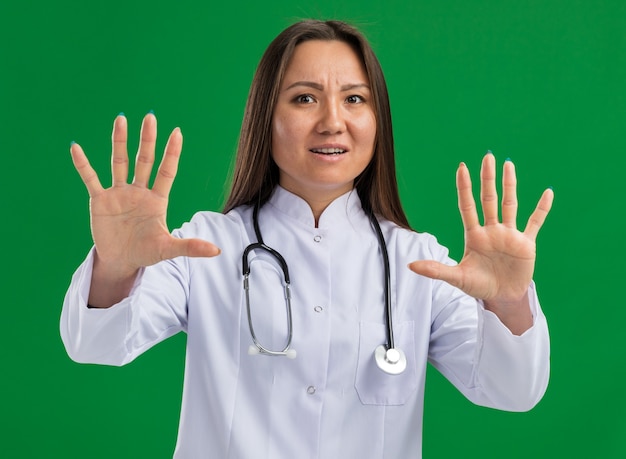 Foto gratuita asustada joven doctora asiática vistiendo bata médica y estetoscopio mirando al frente haciendo gesto de rechazo aislado en la pared verde