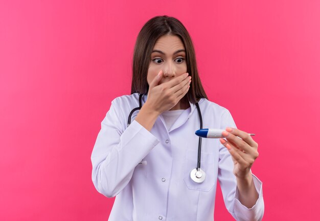 Asustada joven médico con estetoscopio bata médica mirando el termómetro en la mano sobre fondo rosa aislado