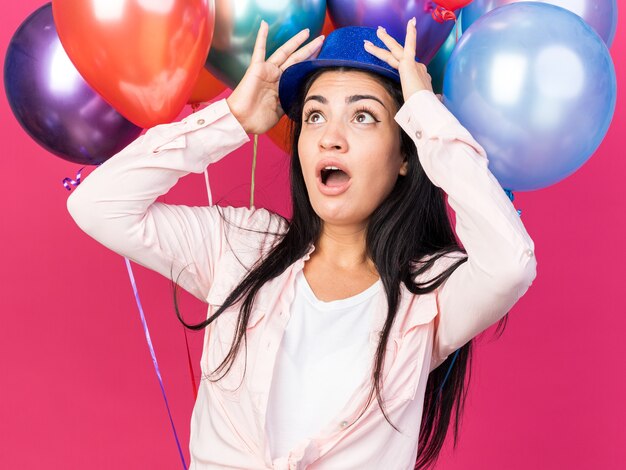 Asustada mirando hacia arriba joven hermosa chica con sombrero de fiesta de pie delante de globos