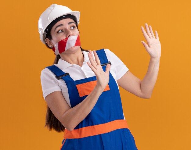 Asustada mirando a la mujer joven constructor de lado en la boca sellada uniforme con cinta adhesiva que muestra el gesto de parada aislado en la pared naranja