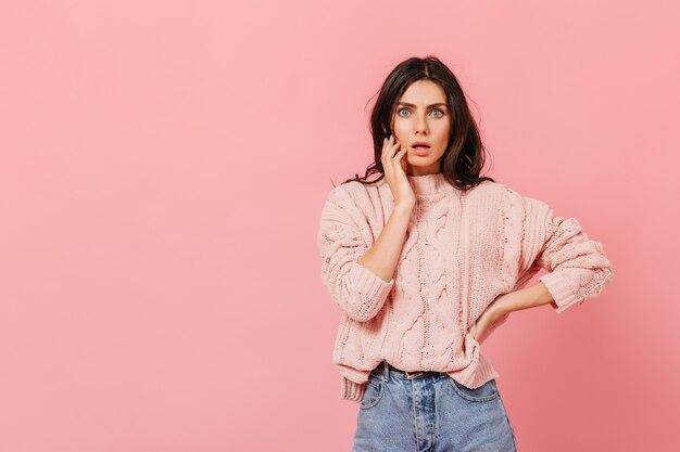 Asustada niña de ojos azules sorprendida mira a la cámara. Instantánea de morena en suéter de punto y jeans en estudio rosa.