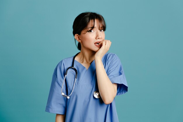 Asustado agarró la barbilla joven doctora vistiendo uniforme fith estetoscopio aislado sobre fondo azul.