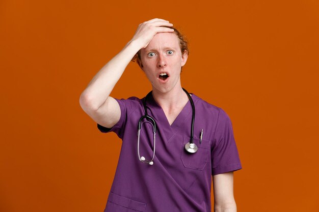 Asustado agarró la frente joven médico masculino vistiendo uniforme con estetoscopio aislado sobre fondo naranja