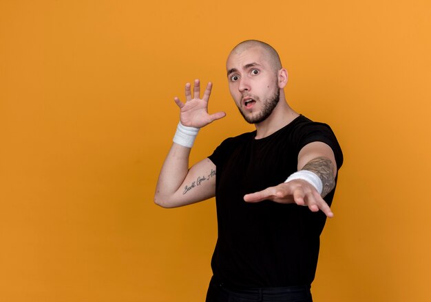 Asustado joven deportivo vistiendo muñequera sosteniendo la mano aislada en la pared naranja con espacio de copia