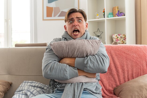 Asustado joven enfermo con bufanda alrededor del cuello abrazando la almohada sentado en el sofá en la sala de estar