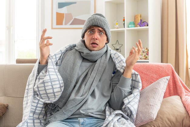 Asustado joven enfermo con bufanda alrededor del cuello vistiendo gorro de invierno envuelto en plaid sentado con las manos levantadas en el sofá en la sala de estar